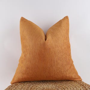 Puede incluir: Un cojín decorativo grande y cuadrado, de color naranja, con una superficie texturizada y acanalada. El cojín descansa sobre una superficie circular tejida. El color del cojín es un tono cálido y acogedor, adecuado para la decoración del hogar.