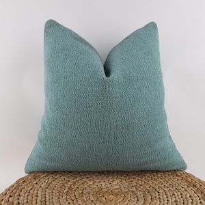 Könnte beinhalten: Ein teal-farbenes Zierkissen mit strukturiertem, gewebtem Aussehen. Das quadratische Kissen liegt auf einer gewebten, kreisförmigen Oberfläche. Der Stoff des Kissens hat eine leicht erhabene, noppige Textur.