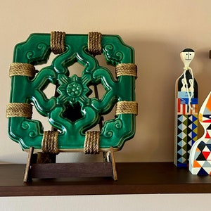 May include: A green ceramic tile with a decorative design, on a wooden easel stand. The tile has a geometric pattern with a central floral motif. Three wooden figurines with geometric patterns stand on a shelf below the tile.