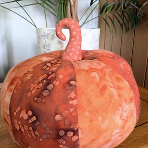 May include: A stuffed fabric pumpkin featuring alternating panels of orange and dark rust-coloured fabric with a mottled, batik-style pattern. The pumpkin has a curved, upright stem made from a lighter orange fabric with a small white floral print.