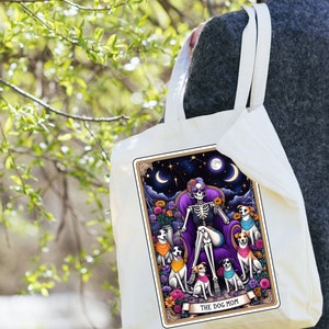 K&ouml;nnte beinhalten: Eine wei&szlig;e Einkaufstasche mit einer bunten Illustration eines Skeletts mit Sonnenbrille, das auf einem Thron mit f&uuml;nf Hunden sitzt. Der Text "THE DOG MOM" ist auf der Tasche aufgedruckt.