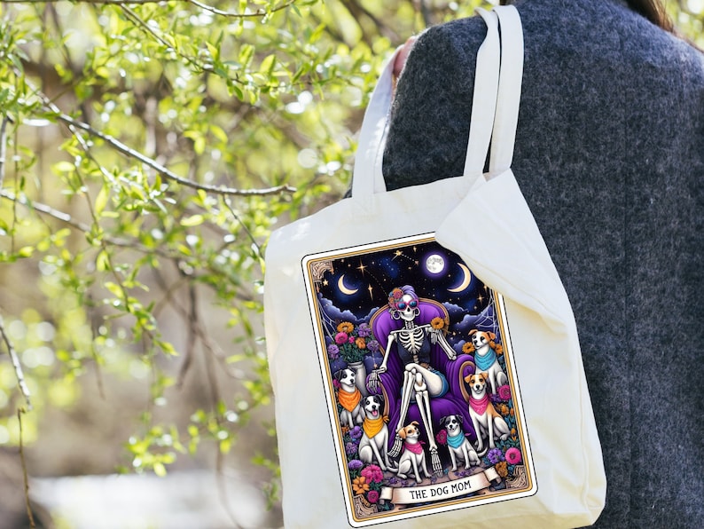 K&ouml;nnte beinhalten: Eine wei&szlig;e Einkaufstasche mit einer bunten Illustration eines Skeletts mit Sonnenbrille, das auf einem Thron mit f&uuml;nf Hunden sitzt. Der Text "THE DOG MOM" ist auf der Tasche aufgedruckt.