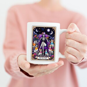 K&ouml;nnte beinhalten: Wei&szlig;e Keramik-Kaffeetasse mit einer farbenfrohen Illustration eines Skeletts mit einer Krone, das auf einem Thron sitzt und von Hunden umgeben ist. Der Text "The Dog Mom" ist am unteren Rand der Illustration aufgedruckt.