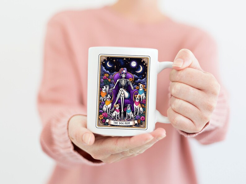 K&ouml;nnte beinhalten: Wei&szlig;e Keramik-Kaffeetasse mit einer farbenfrohen Illustration eines Skeletts mit einer Krone, das auf einem Thron sitzt und von Hunden umgeben ist. Der Text "The Dog Mom" ist am unteren Rand der Illustration aufgedruckt.