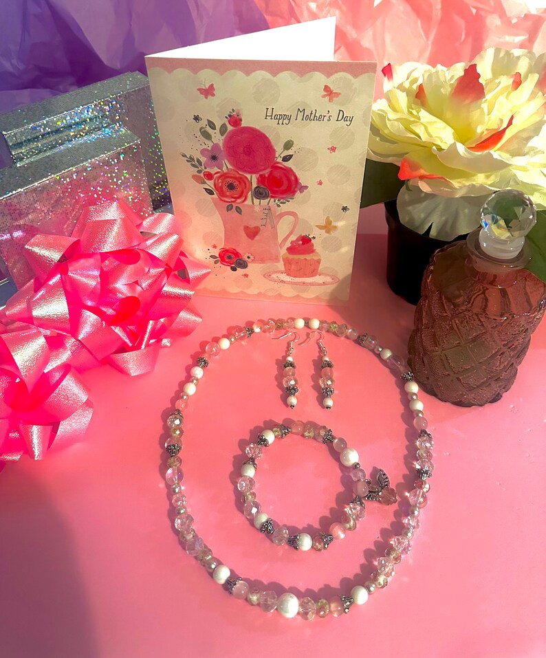 May include: A pink and white jewelry set with a necklace, bracelet, and earrings. The necklace and bracelet feature white and pink beads with silver accents. The earrings are silver with pink and white beads. The jewelry is displayed on a pink surface with a card that says "Happy Mother's Day".