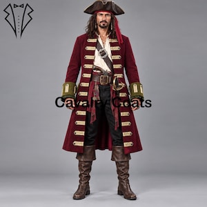 Puede incluir: Un abrigo largo color burdeos con adornos horizontales dorados y puños verde oliva, sobre una camisa blanca y pantalones negros. El conjunto incluye un cinturón de cuero marrón con hebilla, un sombrero de tres picos oscuro y botas altas.