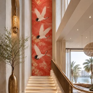 May include: A tall, vertical wall mural featuring three white cranes in flight against a vibrant red background with gold cloud-like accents, positioned above a set of illuminated stairs in a high-ceilinged interior space with a gold floor vase.
