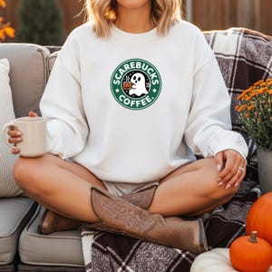 May include: White sweatshirt featuring a Halloween-themed logo that reads "Scarebucks Coffee." The logo includes a ghost holding a coffee cup, surrounded by a green circle with stars. The sweatshirt is a long-sleeved crew neck.