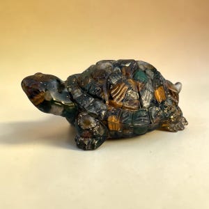 May include: A decorative turtle figurine with a mosaic-like shell. The turtle features a mix of colors, including green, brown, and gold, and is made of a translucent material. The figurine is a detailed representation of a turtle.