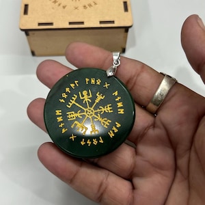 May include: A dark green pendant with gold runic symbols and a silver bail. The round pendant is held in a hand. A wooden box with etched symbols is in the background.