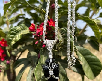 Pode incluir: Um colar prateado com um pingente de pedra preta. O pingente é enrolado em arame e em forma de ponta de flecha. O colar está pendurado em um fundo de folhas verdes e flores vermelhas.