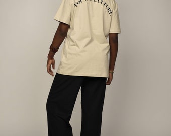Può includere: Una persona indossa una maglietta beige con la scritta "For the Culture" stampata sul retro. La persona indossa pantaloni neri e ha i capelli raccolti in uno chignon.