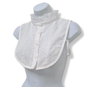 May include: A white, eyelet fabric collar with a frilled high neck and button-down front. The collar is displayed on a mannequin and features delicate floral cutouts and tie closures on the sides.