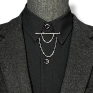 May include: A silver tie bar with two hanging chains is attached to a black dress shirt. The shirt is worn under a dark gray blazer. The tie bar is a stylish accessory for formal wear.