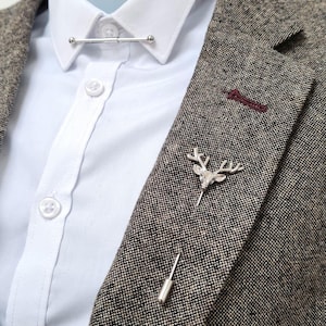May include: A silver collar bar and two lapel pins on a tweed blazer over a white shirt. One pin has a deer head design, the other is a simple, elongated shape. The blazer is a mix of brown and grey.