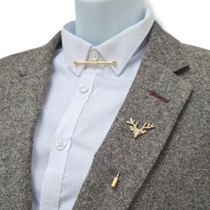 Könnte beinhalten: Ein goldfarbener Krawattenbügel, ein Revers-Anstecker in Form eines Hirschkopfes und eine goldfarbene Anstecknadel schmücken einen grauen Tweed-Blazer über einem hellblauen Hemd. Der Blazer hat einen weinroten Akzent.