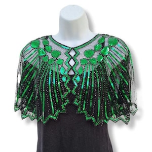 May include: A black mesh capelet adorned with green sequins and beadwork. The capelet has a scalloped hem and a geometric design with green sequins. The garment is displayed on a white mannequin.