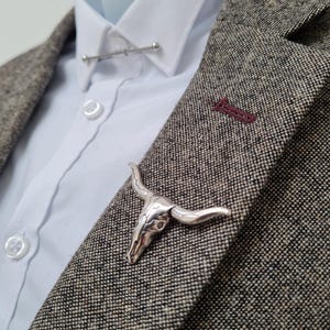 Può includere: Spilla da bavero a forma di teschio di toro argentato e barra per colletto su camicia bianca e blazer in tweed. La spilla a teschio di toro è dettagliata con corna e lineamenti del viso. Il blazer è un mix di toni grigi e marroni.