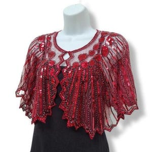 May include: A red sequined capelet with a sheer overlay, displayed on a mannequin. The capelet features a scalloped hem and intricate sequin detailing in a floral pattern. The garment is designed with a keyhole neckline.