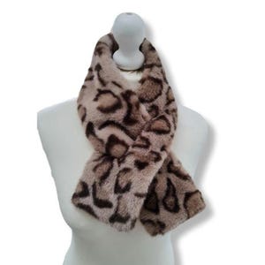 Puede incluir: Bufanda de piel sintética con estampado de leopardo en tonos marrón y beige. La bufanda se muestra sobre un maniquí blanco, destacando su diseño y textura. Un accesorio de moda.