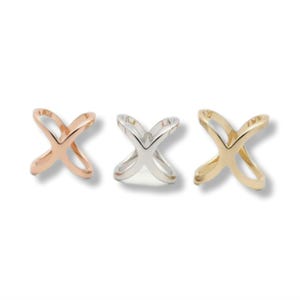 Puede incluir: Tres anillos en forma de X en tonos oro rosa, plata y oro. Los anillos son de metal liso y brillante, presentados sobre un fondo blanco. El diseño es minimalista y moderno.