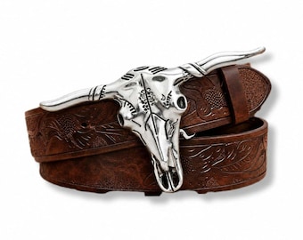 May include: A brown leather belt featuring an embossed floral pattern and a large, silver-toned metal belt buckle shaped like a steer skull with long, curved horns. The belt is coiled, showcasing the detailed texture of the leather and the polished finish of the metallic buckle.