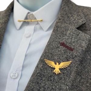 May include: A gold-toned eagle lapel pin attached to a grey textured blazer, paired with a white button-down shirt featuring a gold-toned collar bar pin that connects the two collar points across the front of the shirt.