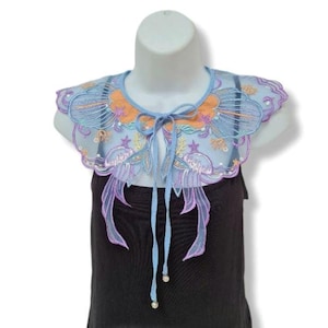 May include: A light blue, embroidered collar with orange and purple floral accents. The collar has a delicate, sheer appearance and is tied with a light blue ribbon. The collar is displayed on a white mannequin head, over a black top.