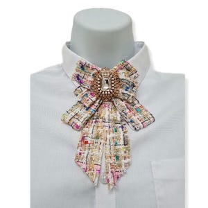 Peut inclure: Un nœud papillon décoratif en tissu façon tweed multicolore, avec des fils roses, bleus et dorés. Le centre est doré, orné d'un grand bijou rectangulaire transparent et de petits bijoux. Le nœud papillon est fixé à une chemise blanche à col.