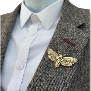 May include: A gold-toned moth brooch featuring a skull pattern on its thorax, pinned to the lapel of a grey tweed blazer worn over a crisp white button-down shirt. The moth has detailed wing textures and is positioned on the left side of the jacket.