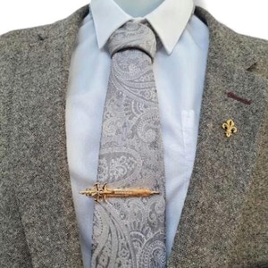May include: A grey paisley tie with a gold tie clip is worn with a light blue shirt and a grey tweed blazer. A gold fleur-de-lis lapel pin is pinned to the blazer.