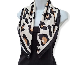 May include: A white and black leopard print silk scarf with a black trim. The scarf is tied around the neck of a mannequin.