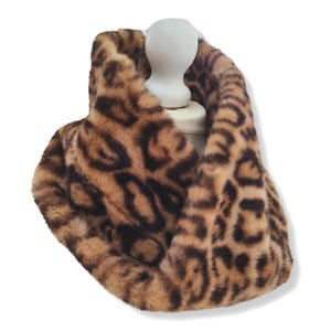 May include: A plush infinity scarf featuring a tan and brown leopard print pattern. The soft, textured fabric is arranged in a circular loop, displaying a repeating animal spot design with dark brown markings against a lighter tan background.