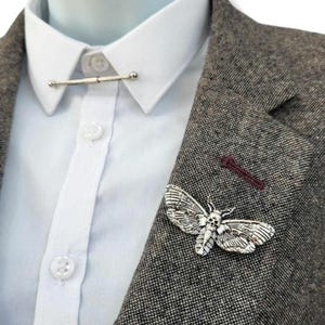 May include: A silver-toned moth brooch with a skull-like pattern on its thorax, pinned to the lapel of a brown tweed blazer. The blazer is worn over a white button-down shirt featuring a silver collar bar connecting the two points of the collar.