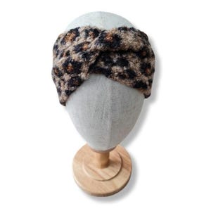 Könnte beinhalten: Ein Stirnband mit Leopardenmuster, das auf einem Schaufensterkopf präsentiert wird. Das Stirnband hat ein braunes, schwarzes und beiges Muster. Der Schaufensterkopf ist hellbeige und auf einem Holzständer montiert.