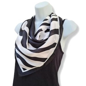 May include: A black and white zebra-print scarf displayed on a mannequin. The square scarf features striking black stripes on a white backdrop. The silky material is draped around the neck, creating a stylish accessory.