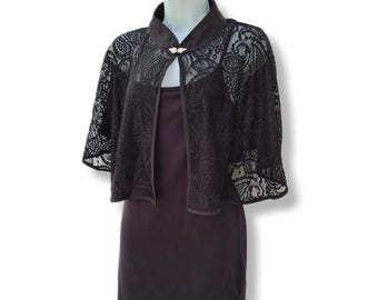 May include: Black lace shrug with a mandarin collar and a button closure. The shrug is worn over a black dress.