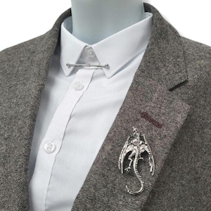 May include: A silver dragon brooch and a silver tie bar on a grey tweed blazer over a white dress shirt. The dragon brooch features outstretched wings and a curled tail. The tie bar is a straight bar with small ball ends.