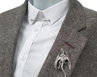 Peut inclure: Broche argentée en forme de dragon et barre de cravate argentée sur un blazer en tweed gris, sur une chemise blanche. La broche dragon a des ailes déployées et une queue enroulée. La barre de cravate est droite.