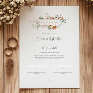 Puede incluir: Un certificado de boda con detalles florales, los nombres "Laura y Martin" y la fecha "06 de junio de 2026" se muestran en papel blanco. Un bolígrafo dorado y dos anillos de oro sobre una superficie de madera.