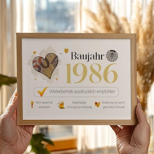 以下が含まれることがあります： 金色の文字で「Baujahr 1986」と書かれた額入りのプリント。プリントには、数字「50」が入ったハートのグラフィック、ダーツボード、その他のアイコンが含まれています。フレームは明るい色の木材で作られています。