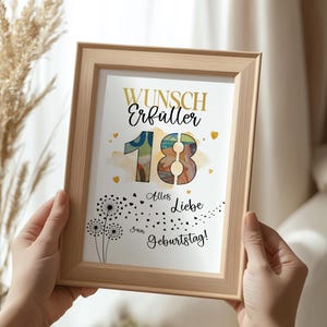 May include: A framed print with the words "Wunsch Erf&uuml;ller 18 Alles Liebe zum Geburtstag!" in gold and black lettering. The number 18 is in a watercolor style. The frame is light brown and is held by two hands.