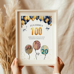 Könnte beinhalten: Eine gerahmte Karte mit einem gold-blauen Ballon-Design. Die Karte sagt "Zusammen 100" und "Herzlichen Glückwunsch zum Doppelgeburtstag!" mit drei Geld-Ballons in der unteren Ecke.