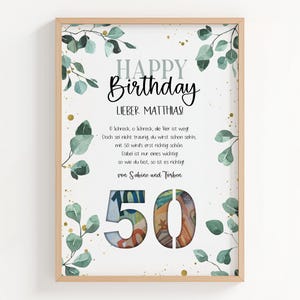 Puede incluir: Una impresión enmarcada con un marco de madera clara. La impresión presenta las palabras "Happy Birthday" y un mensaje en alemán, junto con el número "50" lleno de moneda. Hojas verdes y acentos dorados decoran el borde.