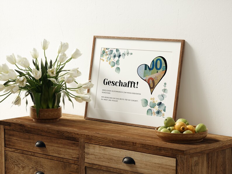May include: A framed print with a watercolor design of eucalyptus leaves and a heart-shaped cutout filled with euro bills. The print has the text "Geschafft!" and a message in German.