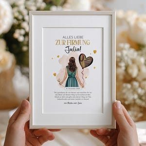 May include: A framed print with the text "ALLES LIEBE ZUR FIRMUNG Julia!" in gold lettering. The illustration features a woman in a teal dress holding a heart-shaped balloon. The print includes the date "16. November 2023" and a short message.