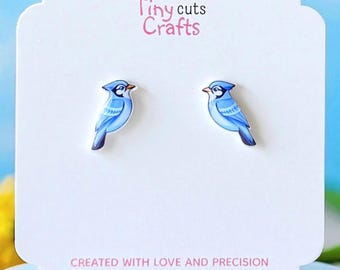 Könnte beinhalten: Ohrringe mit blauen Vögeln auf einer weißen Karte mit dem Text "Tiny cuts Crafts" und "CREATED WITH LOVE AND PRECISION". Die Ohrringe sind vor einem blauen Hintergrund mit rosa und gelben Blumen platziert.
