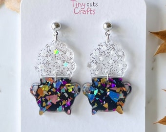 May include: A pair of black and iridescent glitter cauldron earrings with a clear glitter top. The earrings are on a white card with the text "Tiny cuts Crafts" and "Created with love and precision".