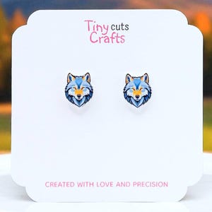 May include: A pair of wolf-themed stud earrings displayed on a white card. The earrings feature a blue and yellow wolf face design. The card has the text "Tiny cuts Crafts" and "CREATED WITH LOVE AND PRECISION".