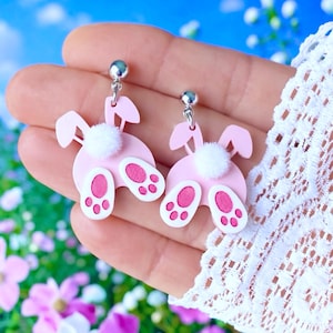 May include: Pink bunny butt earrings with white fluffy tails and pink paw prints. The earrings have silver-colored posts. The background is a blurred image of flowers and a blue sky.